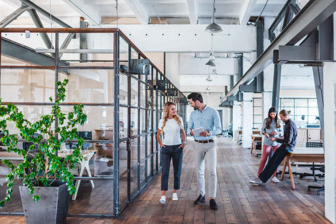 Two professionals walking and talking in a bright, modern open office with glass walls and wooden flooring, while two colleagues collaborate in the background.